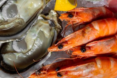 Sashimi çeşnili ton balığı, somon, istiridye, haşlanmış karides. Sashimi büyük, seramik ve hafif bir tabağın üzerinde istiridye kabuğu şeklinde yatar, ortada yaban mersini ve unagi soslu metal kaseler ve avokado tartartarları bulunur. Tabak Stan.