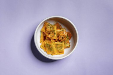 Packeri pasta with shrimp. Wide pasta with chili-tomato sauce, grilled shrimp and finely chopped green onions. Pasta with shrimp lies in a deep, light ceramic bowl. The bowl stands on a purple paper background.