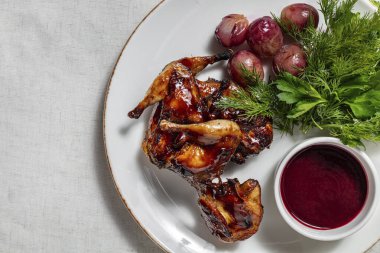 Kiraz soslu ızgara bıldırcın, kiraz soslu üzüm, bir demet maydanoz ve dereotu ve yanında da vişne soslu küçük bir seramik kase. Yemekler yuvarlak, seramik tabakta. Tabak bir ışığın üzerinde duruyor, keten masa örtüsü..