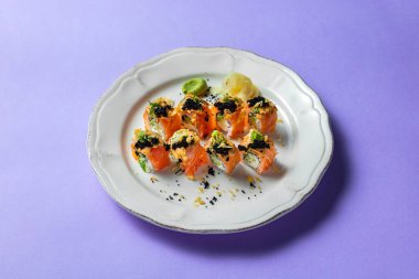 Avokado ve Philadelphia peyniri ile doldurulmuş, somon balığına sarılmış, Japon biberli mayonez ve siyah havyarla kaplı Philadelphia dürümü. Suşi beyaz, yuvarlak, seramik bir tabakta, yanında turşulu zencefil ve bir kare bulunur.