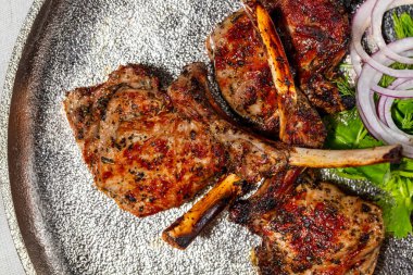 Bir parça dana eti. Kemikte üç parça dana eti var, ızgarada. Parçalar birbirinin üstünde, yanında marul ve kırmızı soğan halkalarından bir yaprak uzanıyor. Kare gümüş, yuvarlak bir tabakta yatıyor. Tabak bir ışıkta duruyor, keten arka plan..