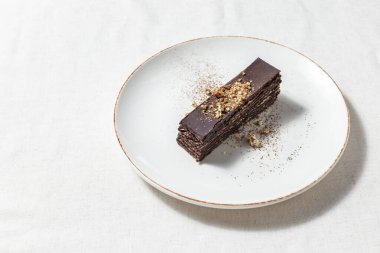 Fındıklı çikolatalı kek. Kakaoya batırılmış fındıklı waffle kekleri. Ezilmiş cevizler pastanın üstüne serpilmiş. Pasta hafif, yuvarlak, seramik bir tabakta yatıyor. Yakınlarda dağılmış bir ceviz var. Tabak bir ışığın üzerinde duruyor, Lin.