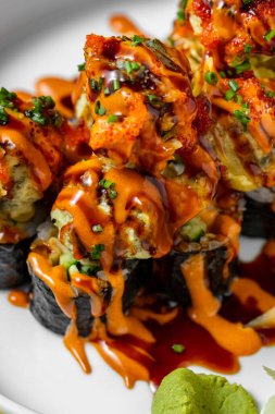 Salmon sushi wrapped in rice and nori seaweed, topped with eel popcorn balls in air dough, unagi sauce and chili sauce, finely chopped green onions are sprinkled on top. Sushi lies on a white, ceramic plate with a pattern. The plate stands on a yello