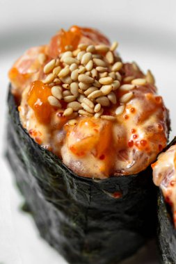 Two gunkans stuffed with rice wrapped in nori and salmon tartare on top. Nunkans stand on a white, round, patterned ceramic plate. Nearby are a square of wasabi and pickled ginger. The plate stands on a purple paper background.
