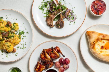 Gürcistan mutfağının çeşitli yemekleri keten masa örtüsünün üzerinde duruyor. Adjarian khachapuri, otlu kuzu pirzolası, kiraz soslu bıldırcın, mantarlı kızarmış patates ve otlu Svan tuzu. Yemek tabaklarının yanında küçük tabaklar var. 