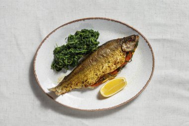 Fırında domates, limon, kişniş ve otlarla pişirilmiş altın nehir alabalığı. Ta ki kahverengiye kadar. Yakınlarda baharatlı ve bir dilim limonlu chukka yosunu salatası var. Yemekler ışık, oval, seramik tabakta. Tabak bir ışığın üzerinde duruyor, keten. 