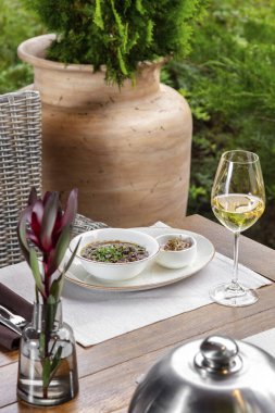 Hafif seramik tabakta soslu ve otlu fasulye lobisi. Onun yanında bir bardak beyaz şarap var. Yiyecek ve şarap ahşap bir masada, hasır bir sandalye ve bitkiler yakınlarda görülebilir..