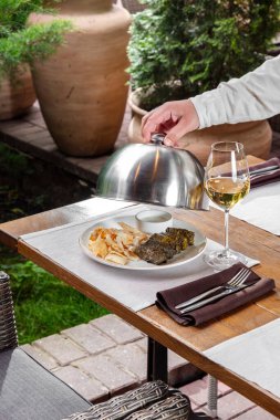 Dolma, yanında lavaş cipsi ve sarımsak sosu bulunan seramik bir tabakta uzanır. Garson kapağı tabağın üzerinde tutuyor, yanında bir bardak beyaz şarap var. Yemek ve şarap ahşap bir masanın üzerinde, hasır bir sandalye ve bitkiler yakında görülebilir.