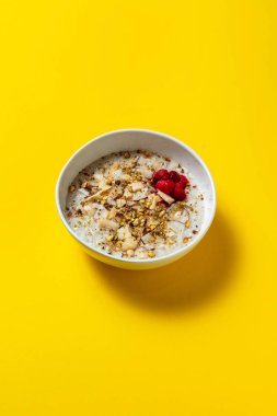 Oatmeal with pistachio, raspberry and coconut in a white plate on a yellow background