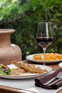 Tavuk omurgası hafif seramik bir tabakta yatıyor, yanında da bir salata var. Tabak ahşap bir masanın üzerinde, yanında bir bardak kırmızı şarap var, sonra bir tabak khachapuri var. Masanın yanında bir koltuk ve bitkiler görülebilir..