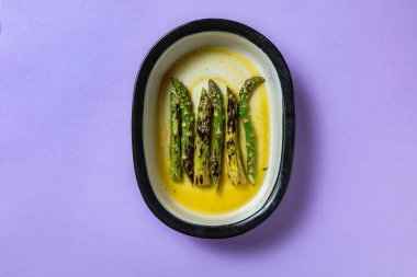 Mor arka planda oval bir tabakta tereyağı ve şarapla kızarmış kuşkonmaz.