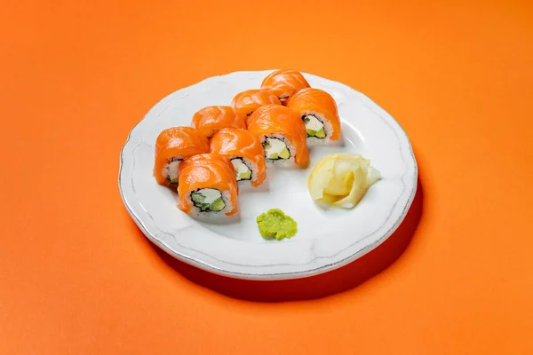 Philadelphia roll with salmon, rice and Philadelphia cheese stuffing with cucumber. Sushi stands on a white, round, ceramic plate with a pattern. The plate stands on an orange paper background.