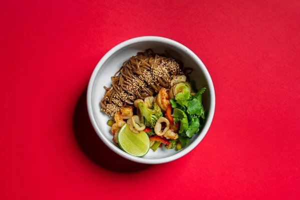 Buckwheat noodles with soy sauce and sesame and squid, shrimp, salmon, parey onion, cilantro leaves, paprika and broccoli florets, half a lime lies nearby. Wok noodles with seafood lie in a deep, light ceramic bowl. The bowl stands on a red paper bac