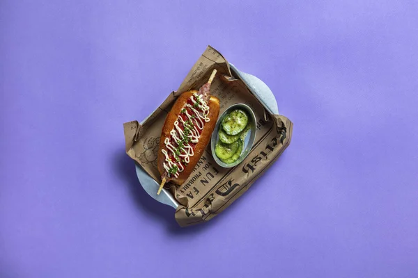 Fish dog with kebab tuna, Japanese mayonnaise and tomato sauce, sprinkled with finely chopped green onions. A fish dog lies on a brown newspaper, next to it is a metal bowl with a pickled cucumber. The food is in a metal tray. Dishes stand on a purpl