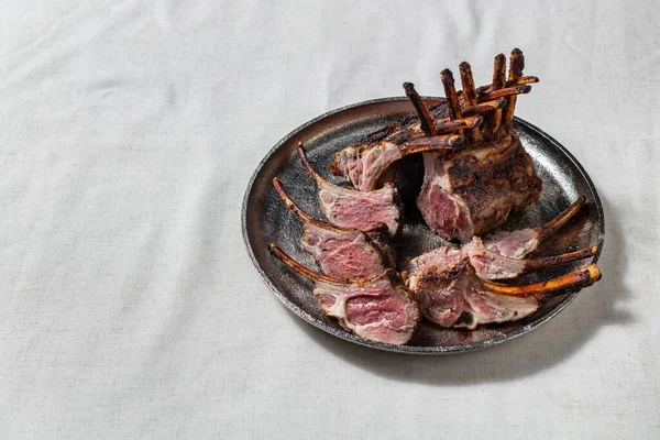 Whole rack of lamb baked in the oven with cherry sauce and herbs. The ribs are partially cut into portions, you can see the degree of roasting and juicy meat. Kare lies on a silver, round tray. The tray stands on a light, linen tablecloth.