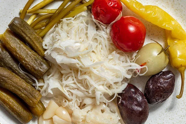 Pickled and salted vegetables. Pickled cucumbers, sauerkraut, pickled garlic, pickled red and green tomatoes, pickled chili peppers, pickled plums and pickled green beans. Vegetables lie on a light, oval, ceramic plate. The plate stands on a light, l