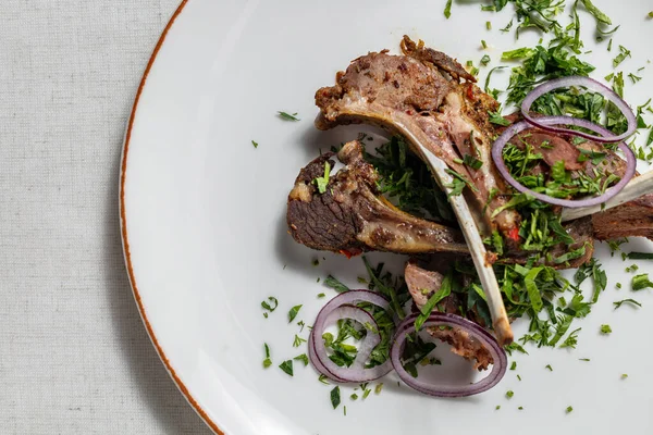 Lamb ribs baked in the oven and cut into portions. Topped with finely chopped cilantro and red onion rings. The ribs lie on a round, light, ceramic plate. The plate stands on a light, linen tablecloth.