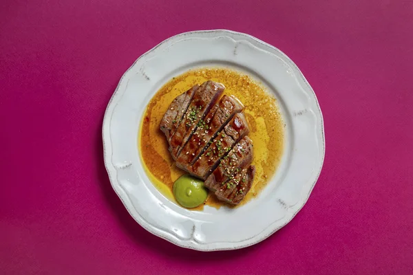 Tuna steak with wine sauce, sesame seeds and green onions in a white plate on a pink background