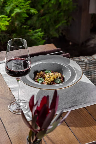 Lamb tartare with spices, herbs, paprika and herbs in a gray, round ceramic plate. The plate is on a wooden table on a stand. Nearby is a glass of red wine. Green plants are visible behind the edge of the table.