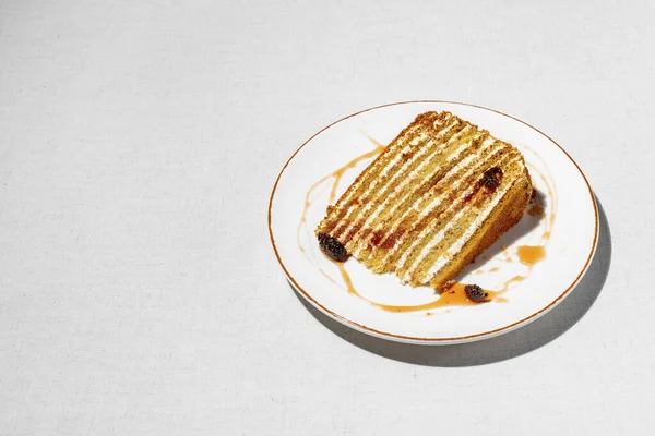 Honey cake. Triangular piece of layer cake Medovik, with cone jam and small cones. The piece lies on a light, round, ceramic plate. Jam is smeared on a plate and cones lie. The plate stands on a light fabric background.