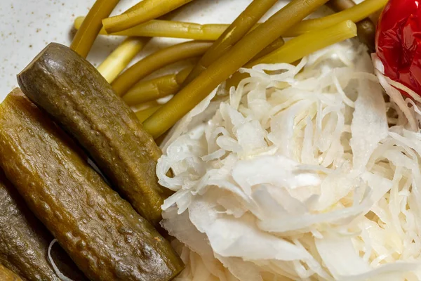 Pickled and salted vegetables. Pickled cucumbers, sauerkraut, pickled garlic, pickled red and green tomatoes, pickled chili peppers, pickled plums and pickled green beans. Vegetables lie on a light, oval, ceramic plate. The plate stands on a light, l