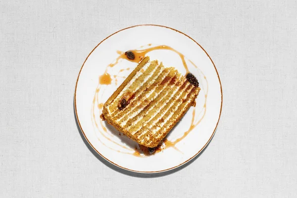 Honey cake. Triangular piece of layer cake Medovik, with cone jam and small cones. The piece lies on a light, round, ceramic plate. Jam is smeared on a plate and cones lie. The plate stands on a light fabric background.