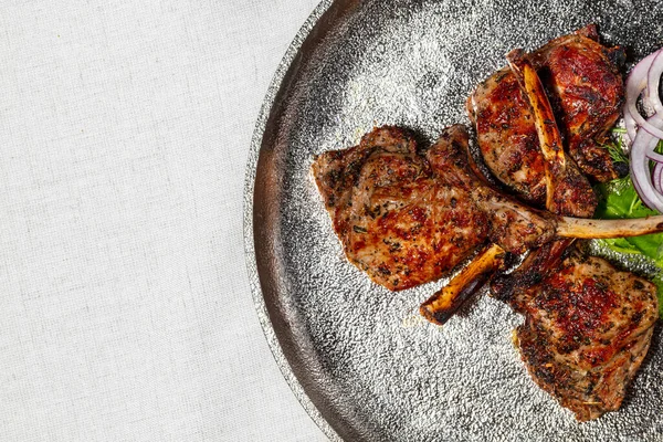 Rack of veal. Three pieces of veal rack on the bone, grilled. The pieces lie one on top of the other, next to it lies a leaf of lettuce and red onion rings. Kare lies on a silver, round plate. The plate stands on a light, linen background.