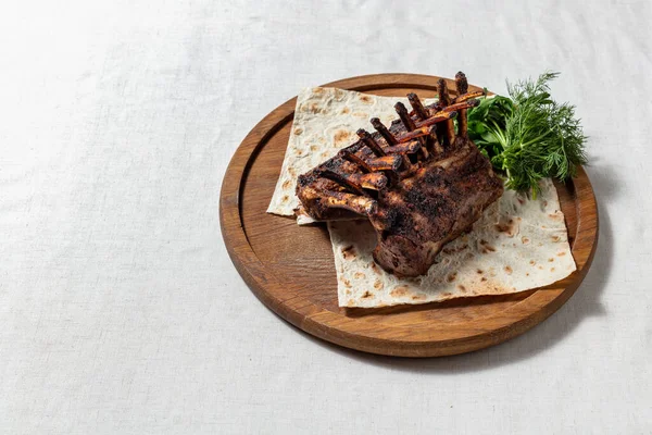 Whole rack of lamb baked in the oven with cherry sauce and herbs. The ribs stand like a house, leaning against each other. on a thin pita bread, next to it lies a bunch of dill and parsley. Food stands on a brown wooden board. The board stands on a l