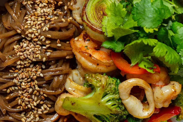 Buckwheat noodles with soy sauce and sesame and squid, shrimp, salmon, parey onion, cilantro leaves, paprika and broccoli florets, half a lime lies nearby. Wok noodles with seafood lie in a deep, light ceramic bowl. The bowl stands on a red paper bac