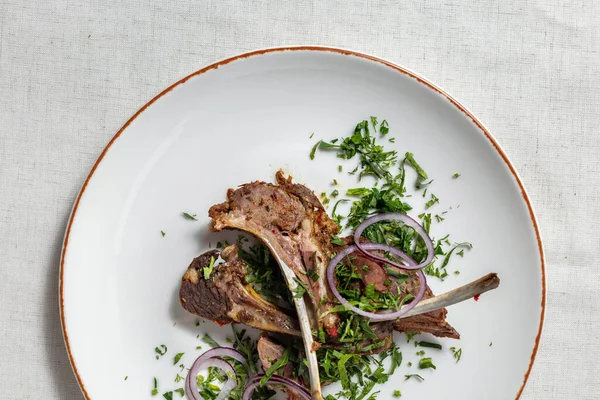 Lamb ribs baked in the oven and cut into portions. Topped with finely chopped cilantro and red onion rings. The ribs lie on a round, light, ceramic plate. The plate stands on a light, linen tablecloth.
