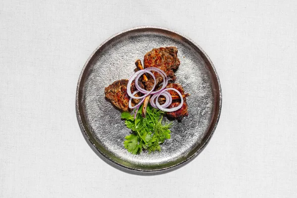 Rack of veal. Three pieces of veal rack on the bone, grilled. The pieces lie one on top of the other, next to it lies a leaf of lettuce and red onion rings. Kare lies on a silver, round plate. The plate stands on a light, linen background.
