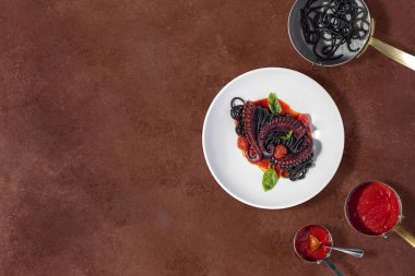 Spaghetti with cuttlefish ink, cherry tomatoes, basil leaves and grilled octopus. The food lies in a light ceramic plate and stands on a brown, stone background. Nearby are saucepans with sauce and pasta.