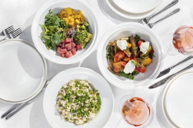 Assorted salads. Olivier salad is placed in a light ceramic dish. tuna salad and baked vegetable salad. Plates stand on a light tablecloth, glasses of wine stand nearby.