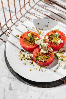 Three round slices of watermelon with feta cheese, nuts and herbs in olive oil. Food lies on a light ceramic plate on a marble countertop. Nearby is a glass of rose wine and a vase of flowers.