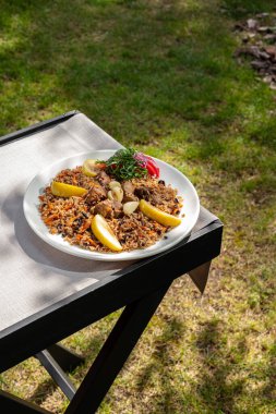 Pilaf with lamb, apples and a mix of fresh vegetable salad on a light ceramic plate. The plate is on a wooden table, next to a glass of white wine. The table is on the lawn.
