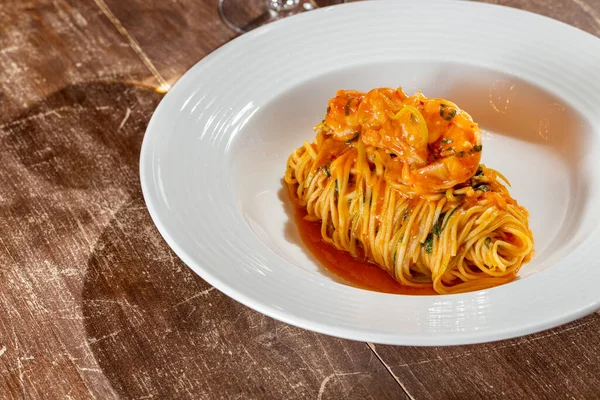 意大利面 番茄酱加罗勒和虾仁 食物放在一个薄薄的陶瓷盘子里 旁边是一杯白葡萄酒 饭菜放在一张木制桌子上 — 图库照片