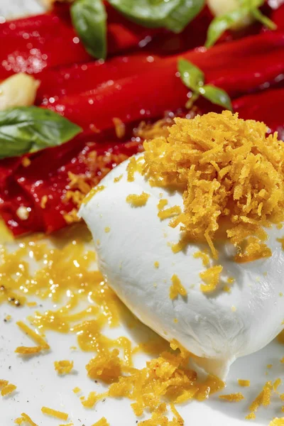 A whole ball of mozzarella with potato chips lies on a light-colored plate, next to a portion of pieces of pickled paprika and garlic with basil leaves. Next to the plate is a glass of rose wine. The plate stands on a light marble countertop.