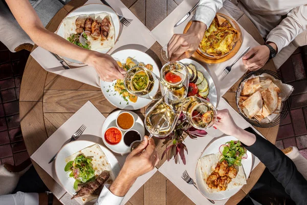 Lunch of chicken and pork skewers, chicken in a creamy sauce, potatoes and grilled vegetables. Side by side are wine glasses and sauces. Food lies on light ceramic plates on a wooden table, people are sitting nearby.