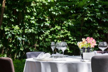 A round table with a white tablecloth stands in the middle of a lawn and a fence of grapes. Plates with underplates are served on the table. cutlery and wine glasses. There are chairs around the table.