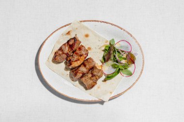 Pork kebab. Four pieces of pork kebab on a sheet of thin Armenian lavash with a mix of greens and radish salad. The food lies on a ceramic plate, on a light tablecloth.