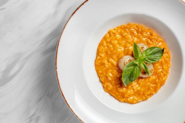 Italian dish risotto with cheddar cheese and grilled scallop, with basil leaves on top. Food in a light ceramic plate with wide margins on a marble background.
