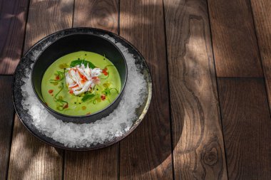 Broccoli and spinach cream soup with crab meat, herbs and balsamic sauce. Soup in a black ceramic plate on ice crumbs in a metal bowl. Dishes stand on a wooden background.