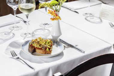 Domates ezmeli karabuğday ekmeği bruschetta, kereviz yaprağı, kapari ve enginar. Hughchetta beyaz bir masa örtüsünün üzerinde açık seramik bir tabağın üzerinde duruyor. Yakınlarda bir bardak beyaz şarap, bir vazo ve birkaç boş bardak var..