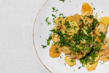 Sliced potatoes and fried in a frying pan with corn oil. Topped with finely chopped parsley. The food lies on a light, ceramic plate on a light tablecloth.