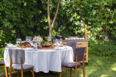 On a round table with a white tablecloth are dishes from the oven and barbecue. Duck with apples, an idea with oranges, lamb ribs, steaks on light ceramic plates. The table stands on a lawn among greenery.