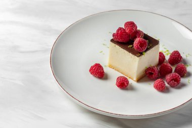 Dessert cheesecake on a nut crust with chocolate icing. Raspberries and lime zest lie on top and next to it. The dessert lies on a light ceramic plate on a marble surface.