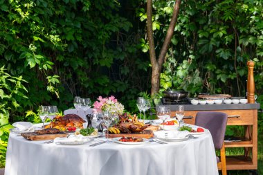 On a round table with a white tablecloth are dishes from the oven and barbecue. Duck with apples, an idea with oranges, lamb ribs, steaks on light ceramic plates. The table stands on a lawn among greenery.
