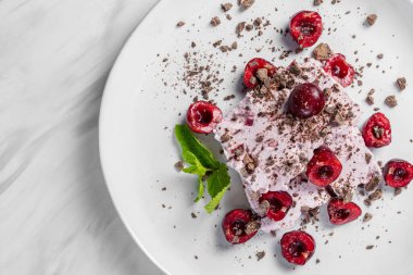 Two slices of creamy cherry ice cream lie on a light ceramic plate. Topped with chocolate chips and cherries with mint leaves. The plate sits on a marble surface.