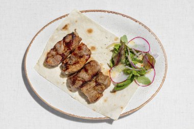 Pork kebab. Four pieces of pork kebab on a sheet of thin Armenian lavash with a mix of greens and radish salad. The food lies on a ceramic plate, on a light tablecloth.