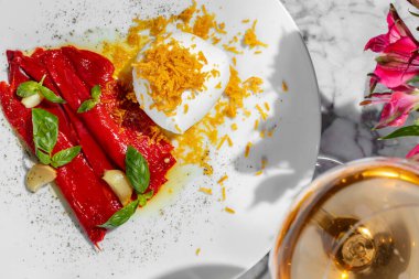 A whole ball of mozzarella with potato chips lies on a light-colored plate, next to a portion of pieces of pickled paprika and garlic with basil leaves. Next to the plate is a glass of rose wine. The plate stands on a light marble countertop.